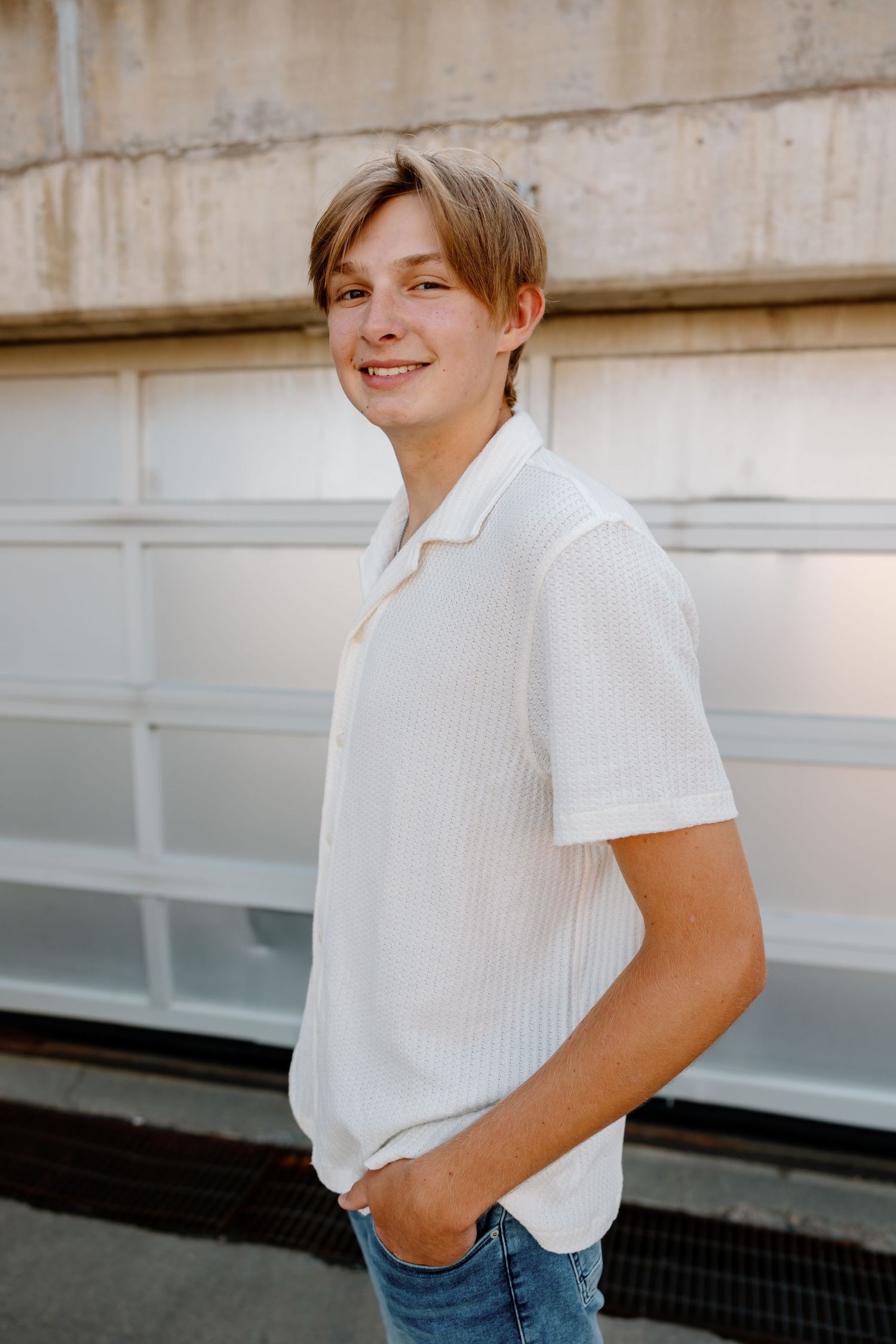 Person wearing white collared shirt and jeans poses against concrete wall outdoors.