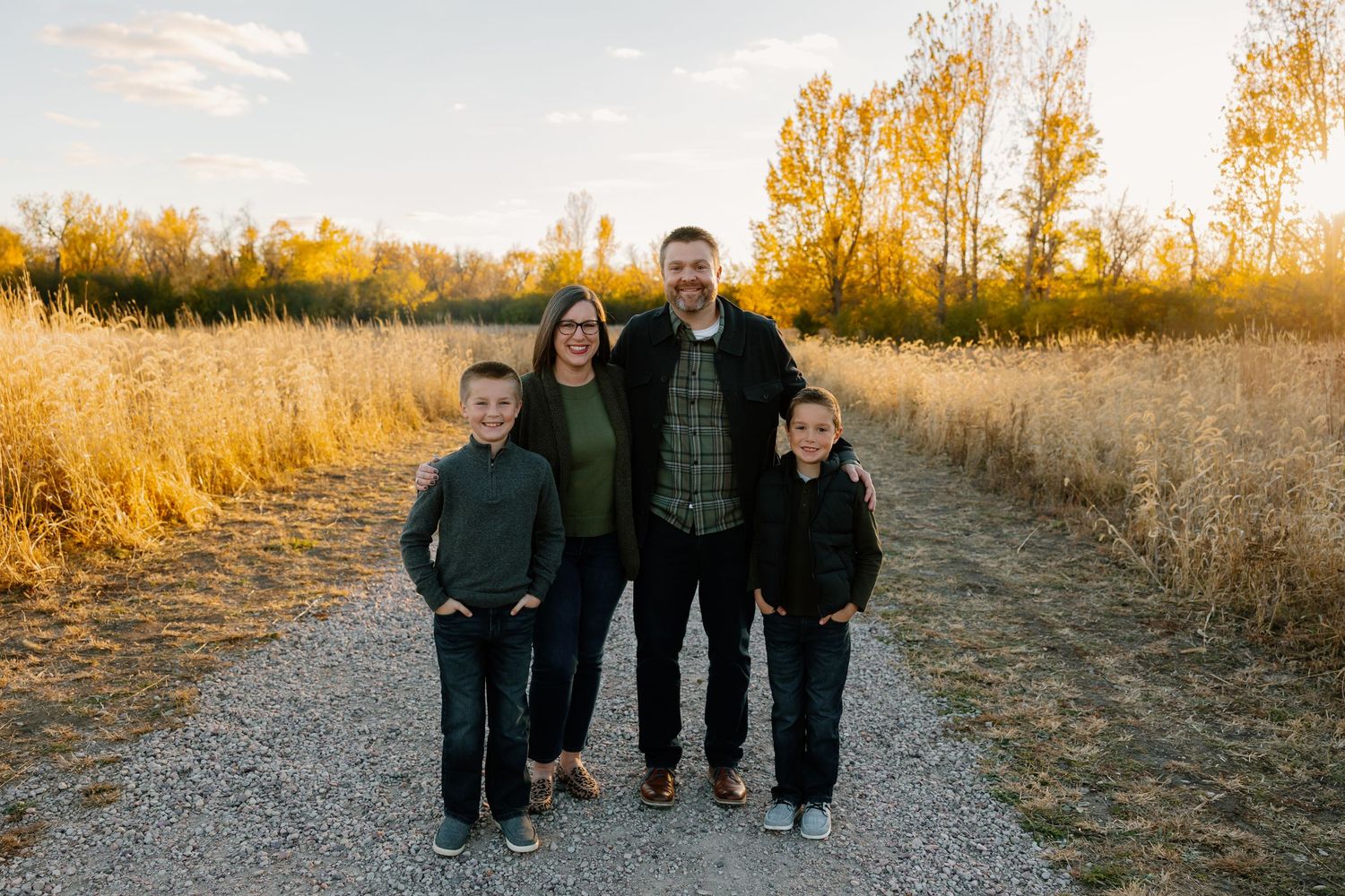 Family of four smiling on a gravel path surrounded by golden autumn trees at sunset.