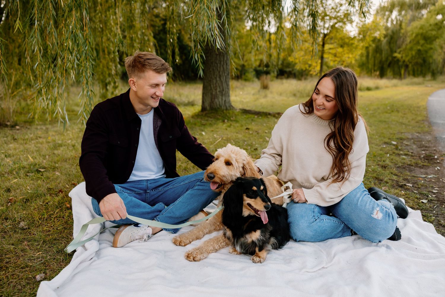 Couple sitting on white blanket in park with two dogs, smiling outdoors near willow tree on autumn day.