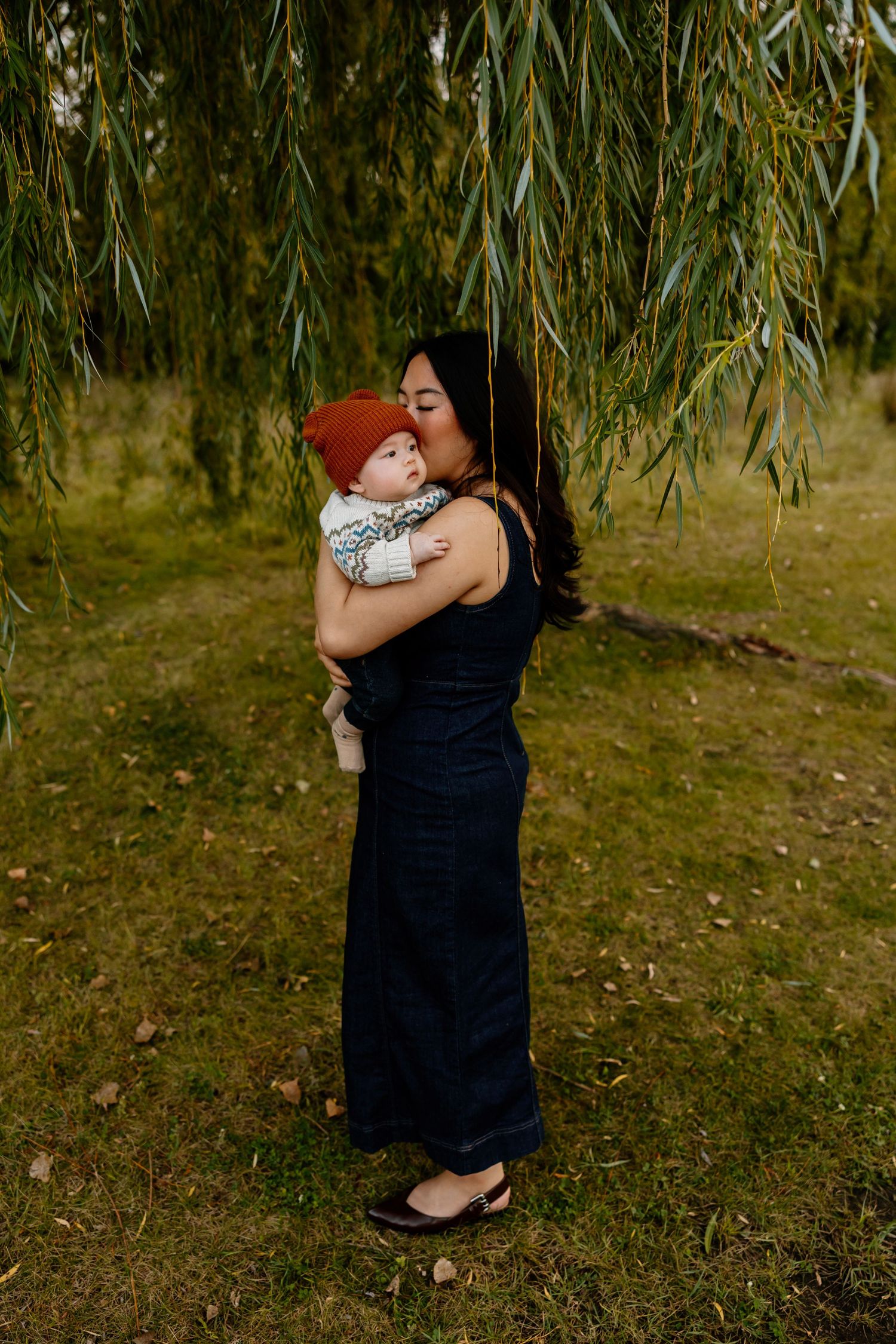 A mother in a navy jumpsuit kisses her baby wearing a red knit hat under a weeping willow tree.