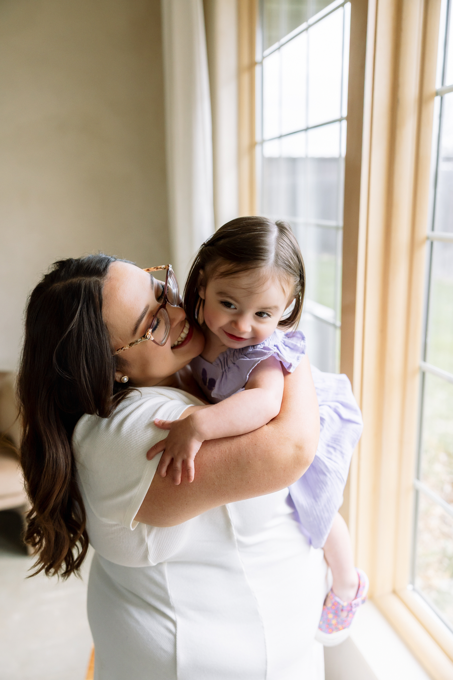 A mother in white holds her smiling toddler girl in a purple dress near a bright window.