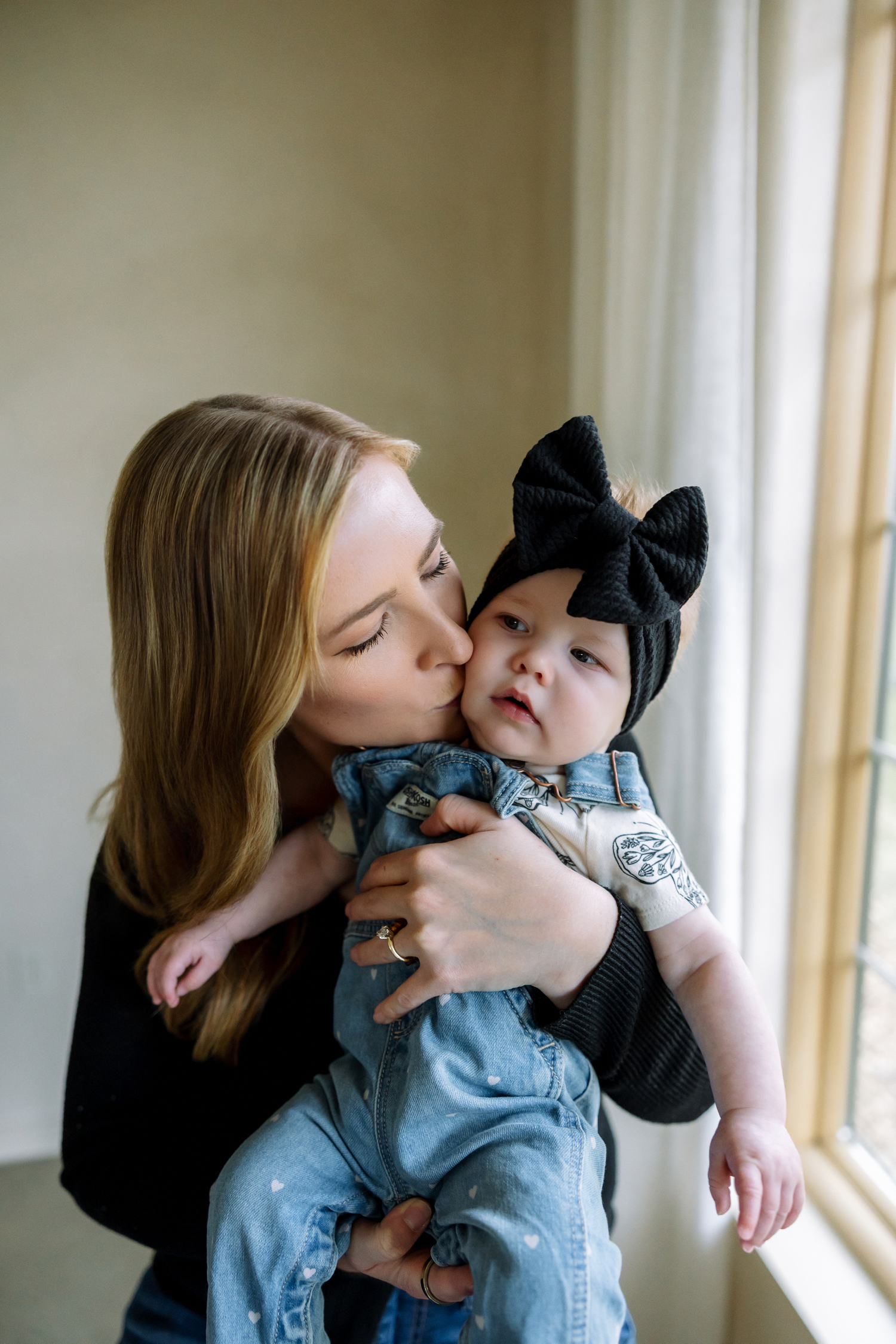 A loving mother kisses her baby girl wearing a black bow headband and denim outfit near a bright window.