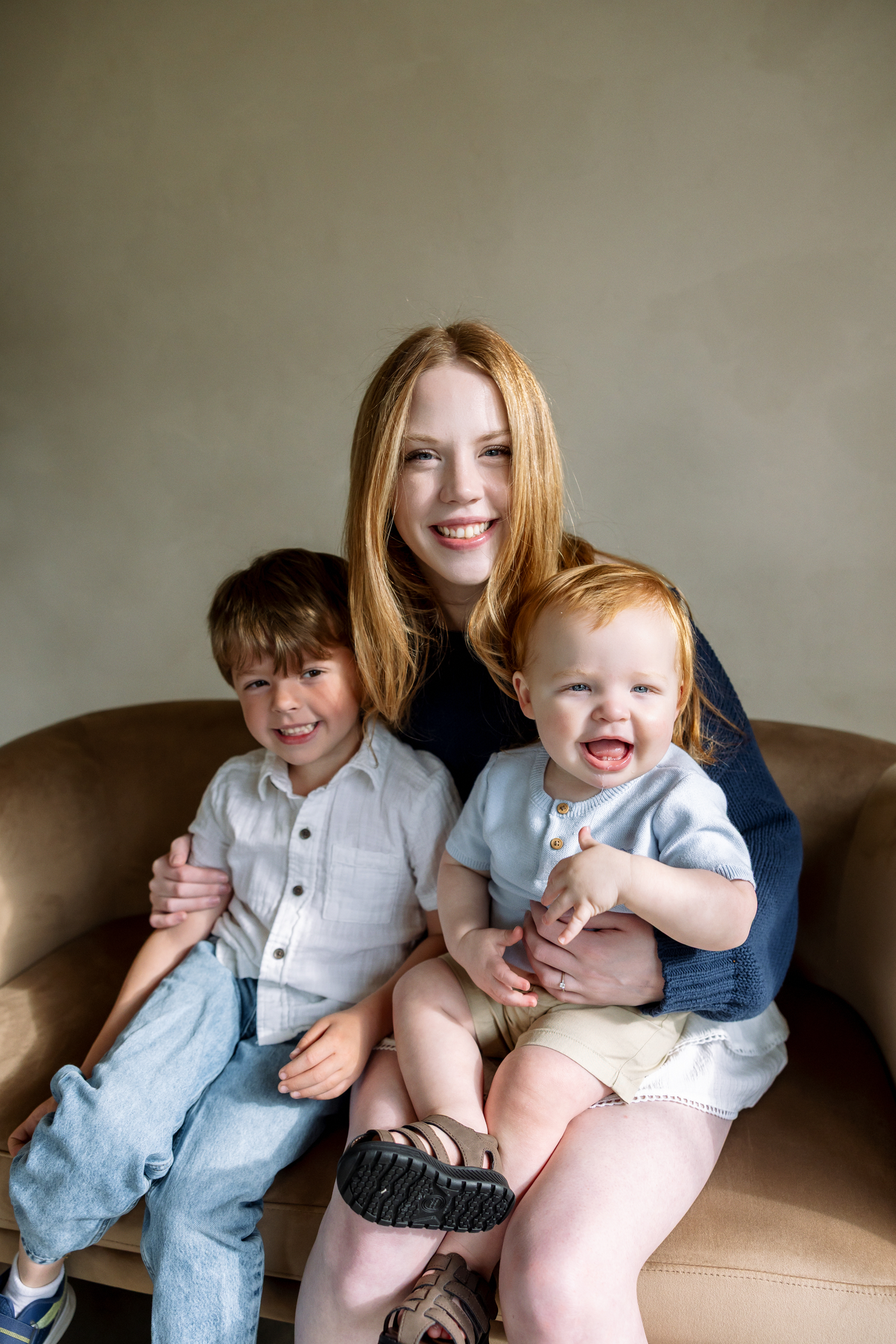 A smiling redhead mother sits on a leather chair with her two young children, a toddler boy and a baby, all dressed in blue.