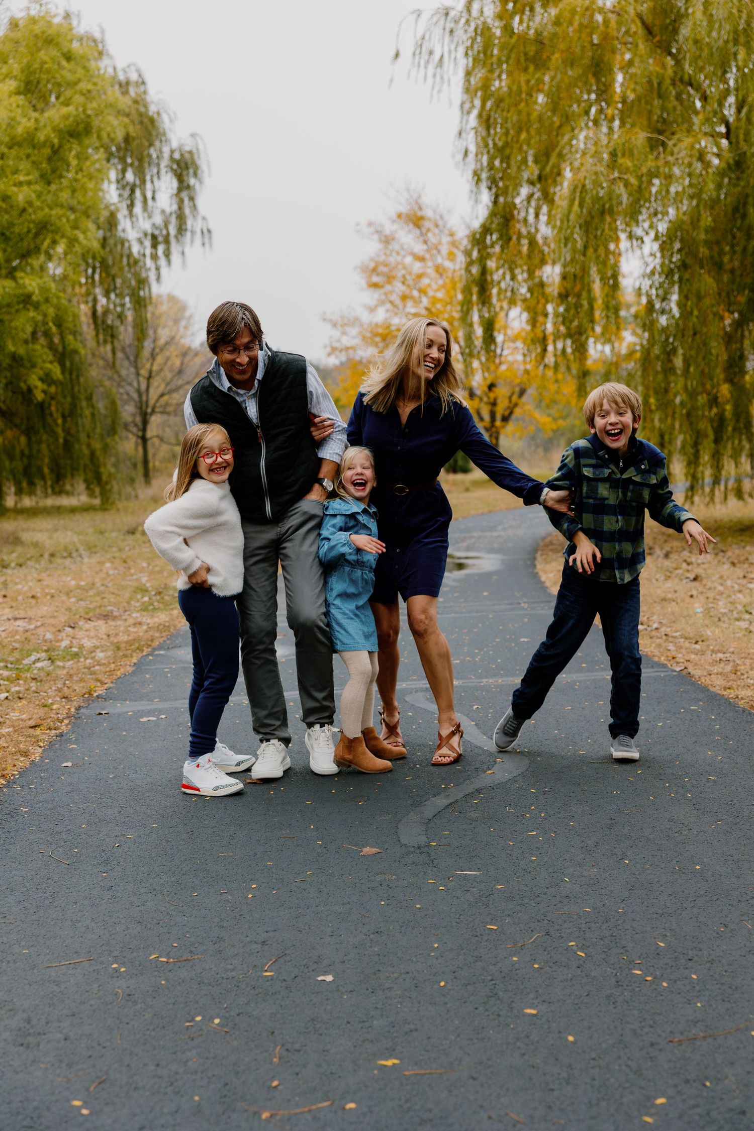 A sequence of playful family photos showing people sitting and posing together on a paved path surrounded by autumn trees.