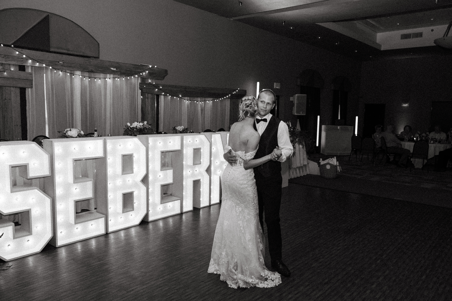 Bride and groom sharing first dance in front of illuminated SEBERT marquee letters at wedding reception.