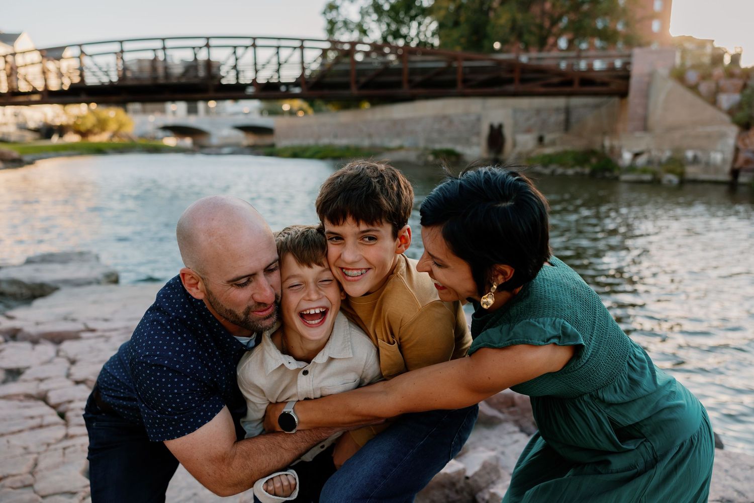 A joyful family of four laughs together by a scenic riverside, with a bridge in the background at golden hour.