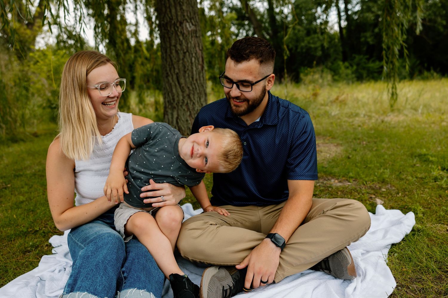 A family sits together on a white blanket in a sunny park, sharing a playful moment outdoors.