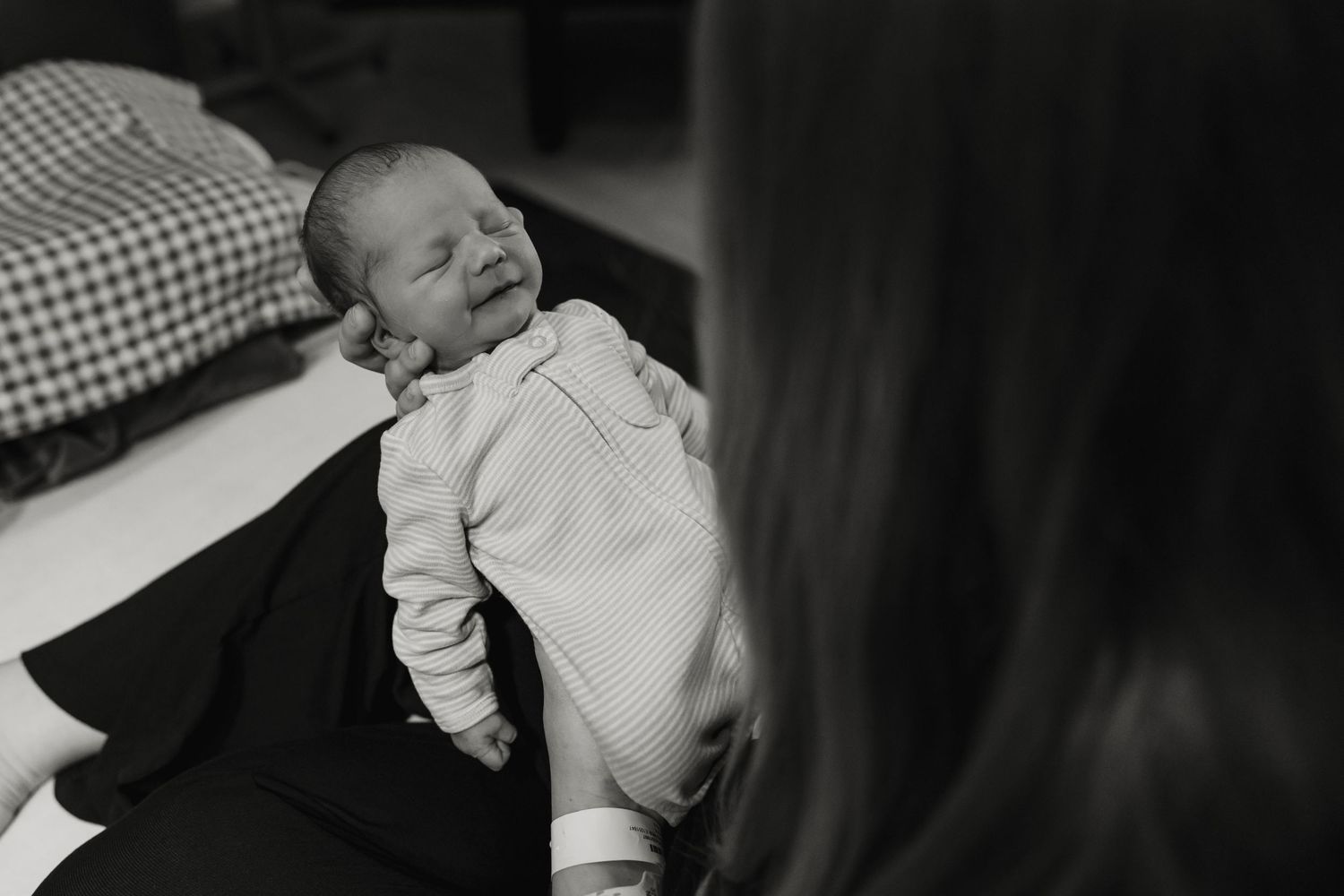 A black and white photo captures a newborn baby smiling while lying on dark fabric.