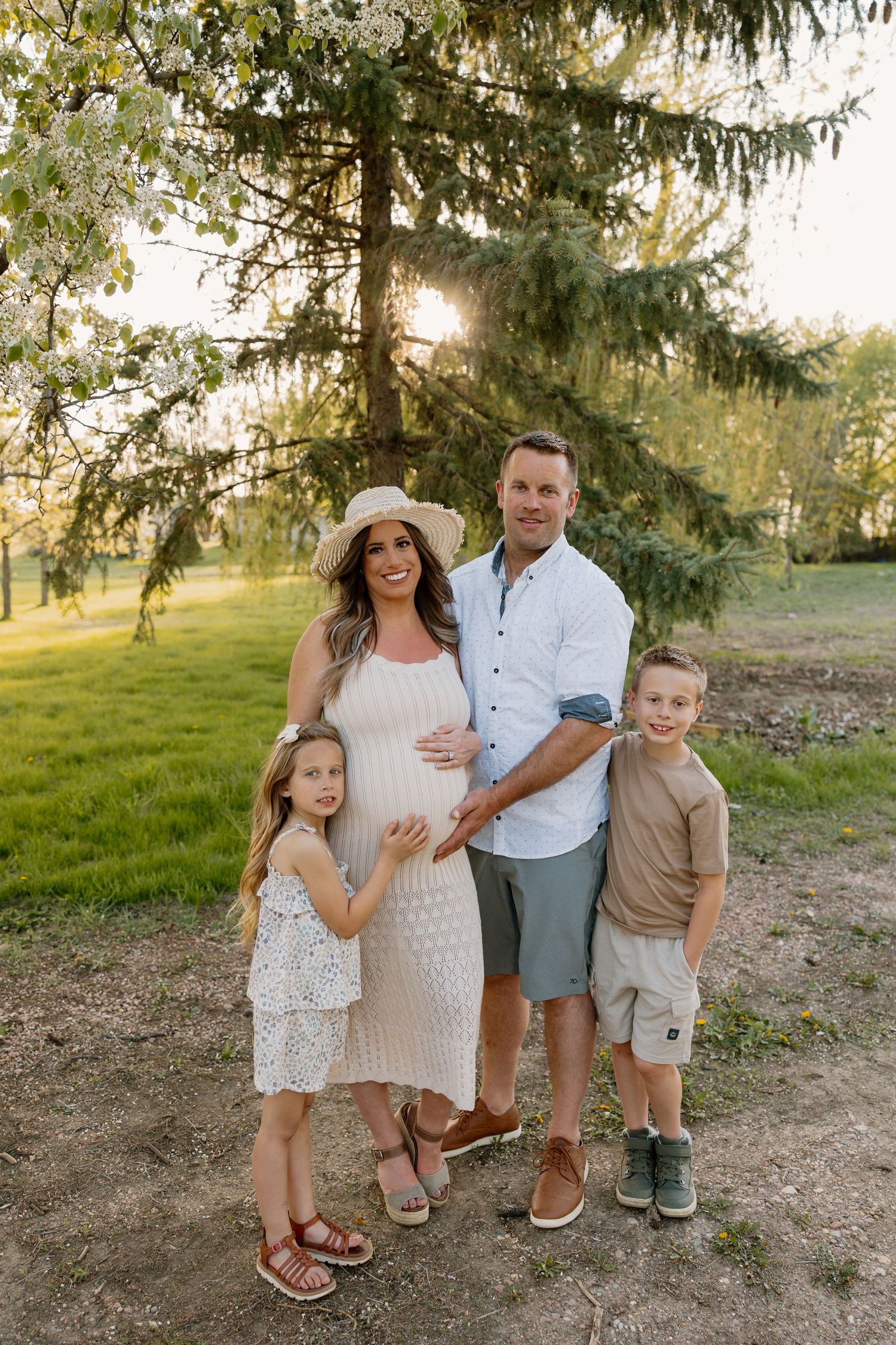 A family poses together outdoors under pine trees at sunset, wearing casual summer clothing.