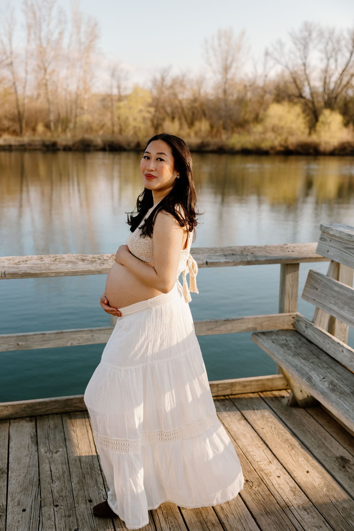 Person in a flowing white dress poses on a wooden dock by a tranquil lake during golden hour.
