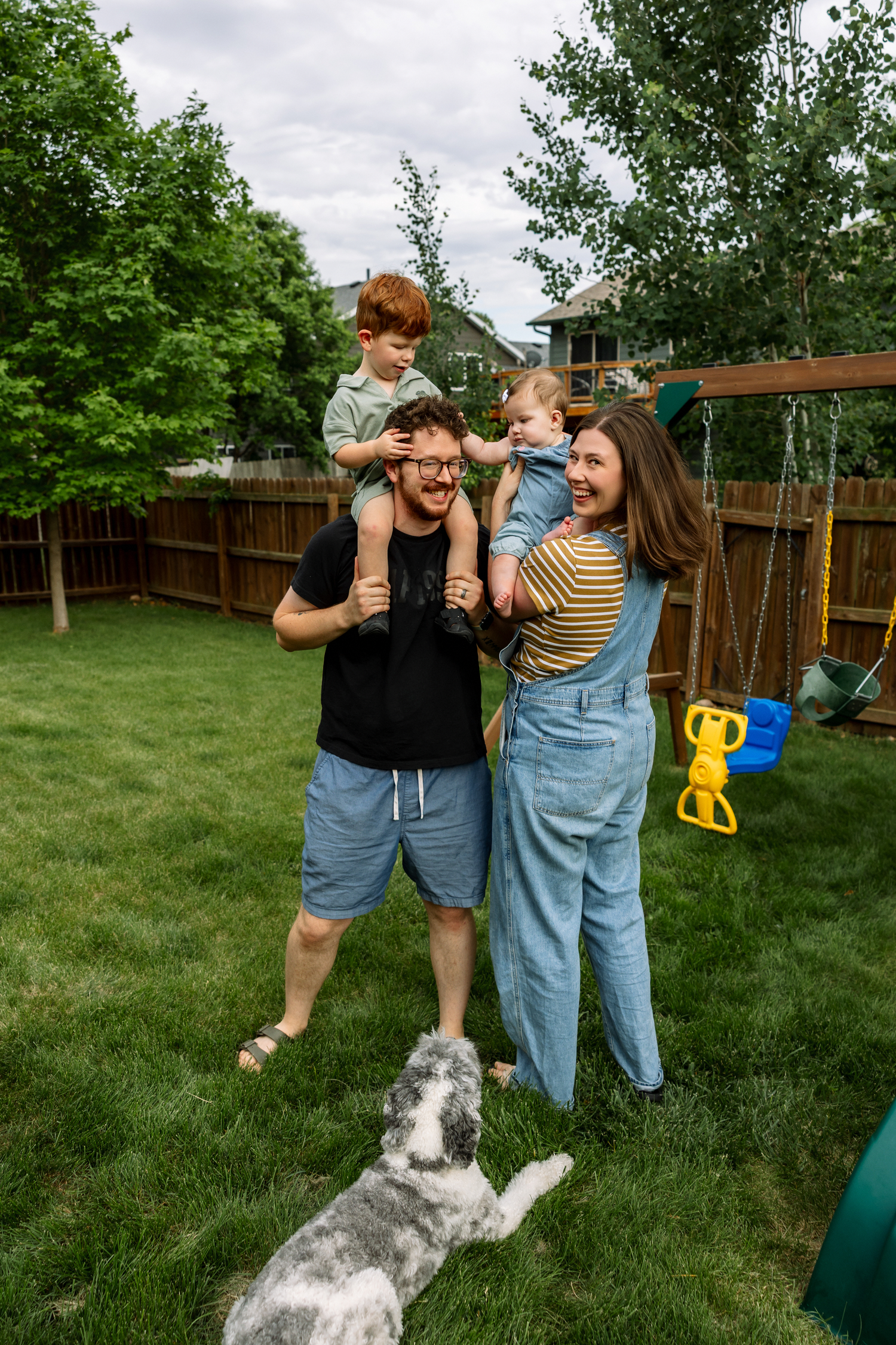 Happy family with two young children and dog playing in green backyard near wooden swing set.