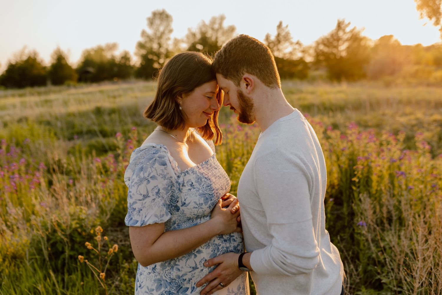 Pregnant couple holding hands in golden sunset field, sharing tender moment surrounded by wildflowers.