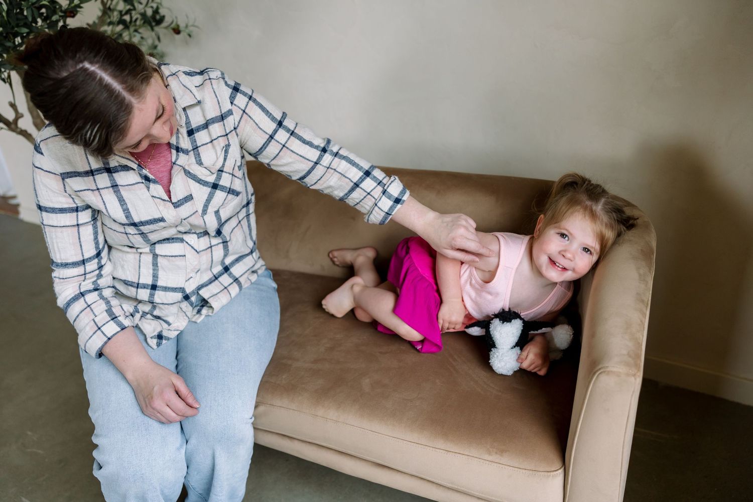 A mother playfully tickles her laughing toddler girl in a pink outfit on a beige couch indoors.
