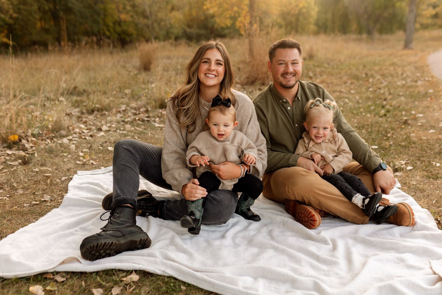 Happy family of four sitting on white blanket in autumn field, smiling outdoors together.