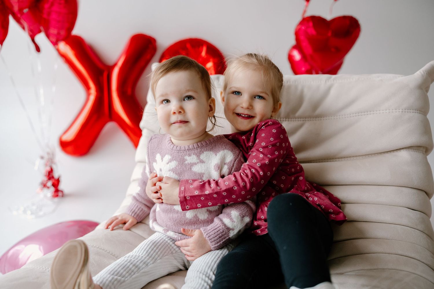 Two children in festive Valentine's Day attire sit on white couch with red heart decorations in background.