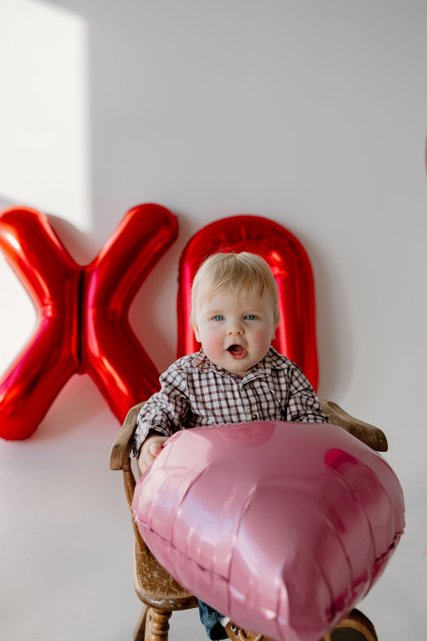 Small child in plaid shirt sits with heart-shaped balloon decoration and red XO lettering against white wall.