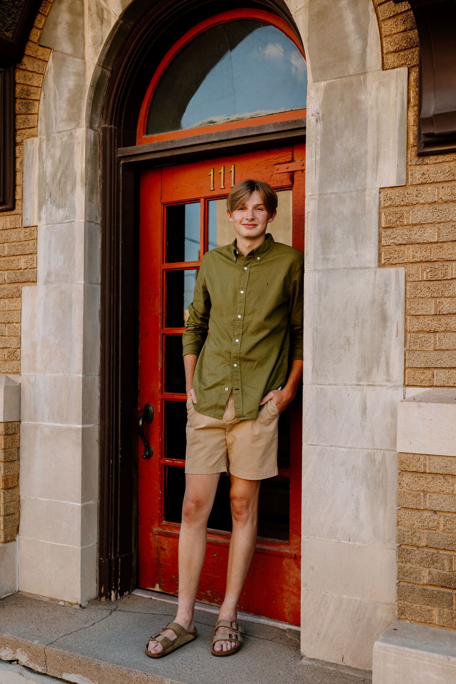 Person in an olive green button-down shirt poses playfully by stone columns and red doors.