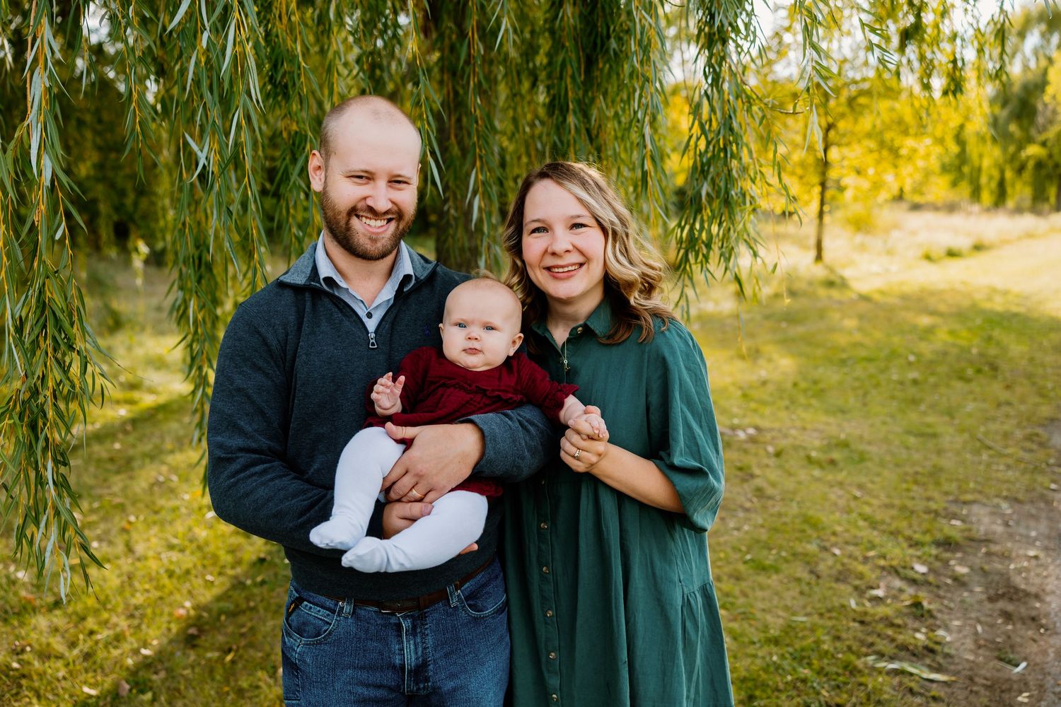 Happy young family with baby posing under willow tree in autumn park setting.