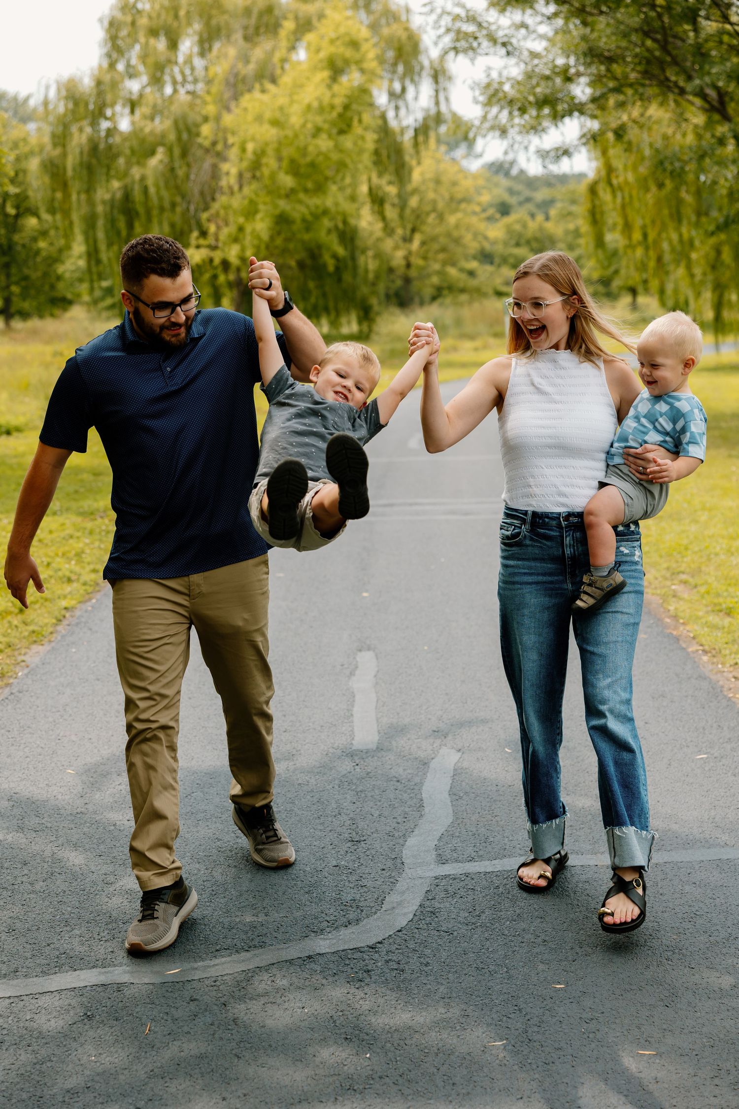 Family enjoying a playful moment together on a tree-lined path, swinging child while walking outdoors.