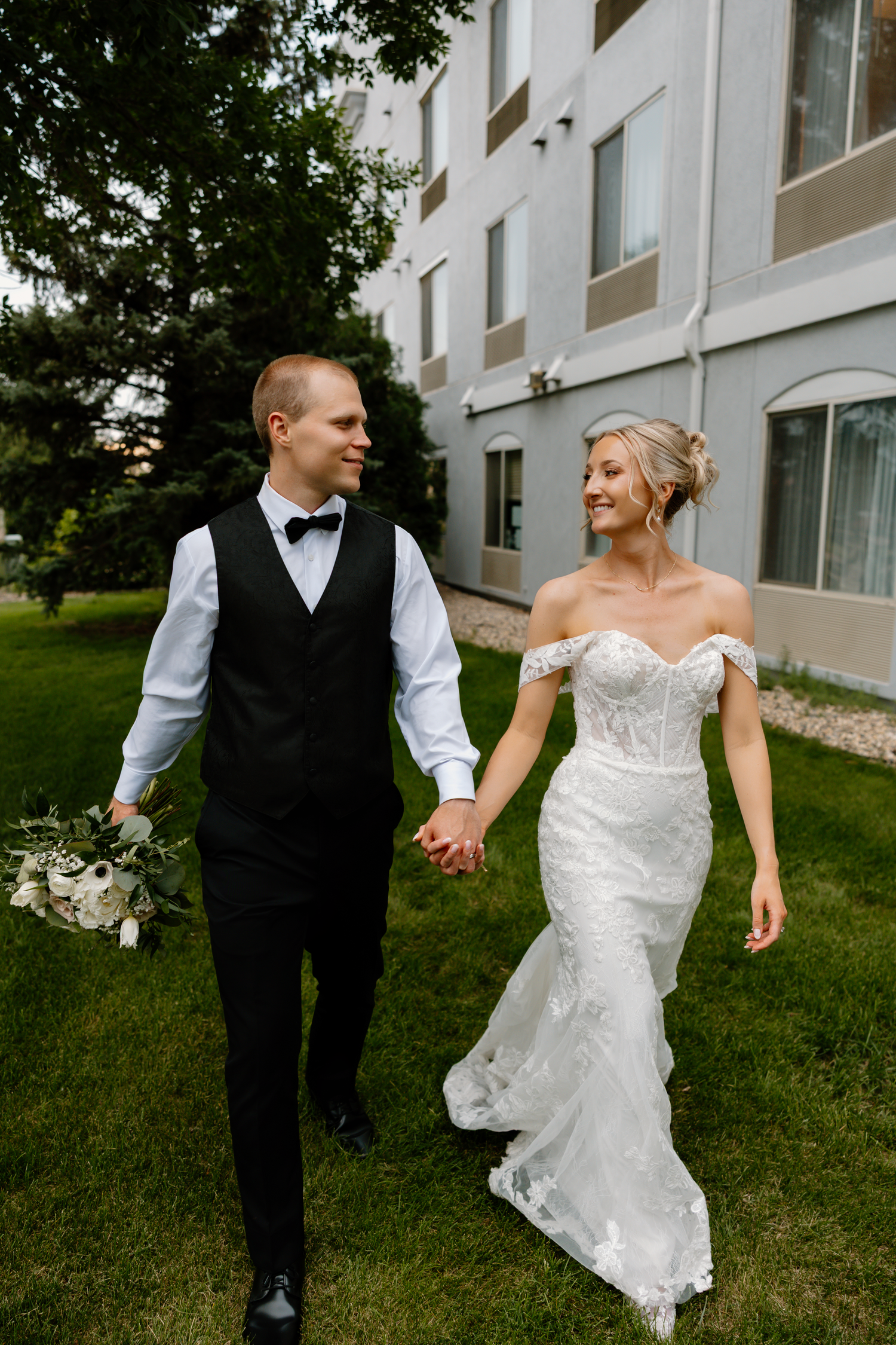 Bride in lace mermaid gown and groom in black vest walk hand in hand on green lawn.