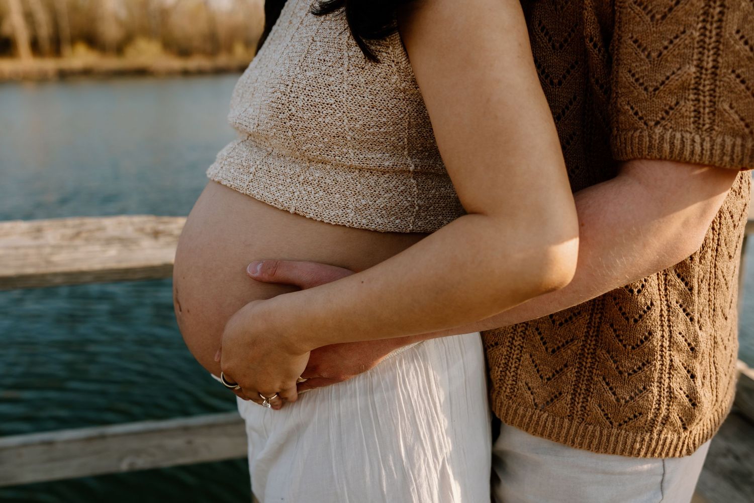 A pregnant person in a cream-colored top stands near a lake at sunset while cradling their baby bump.