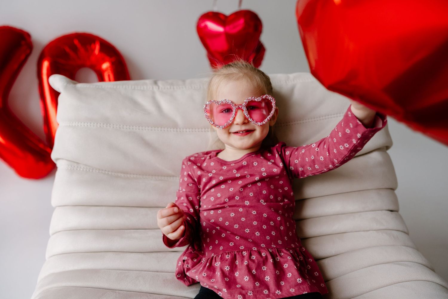Child in pink polka dot dress and heart sunglasses poses on white couch with red heart decorations.