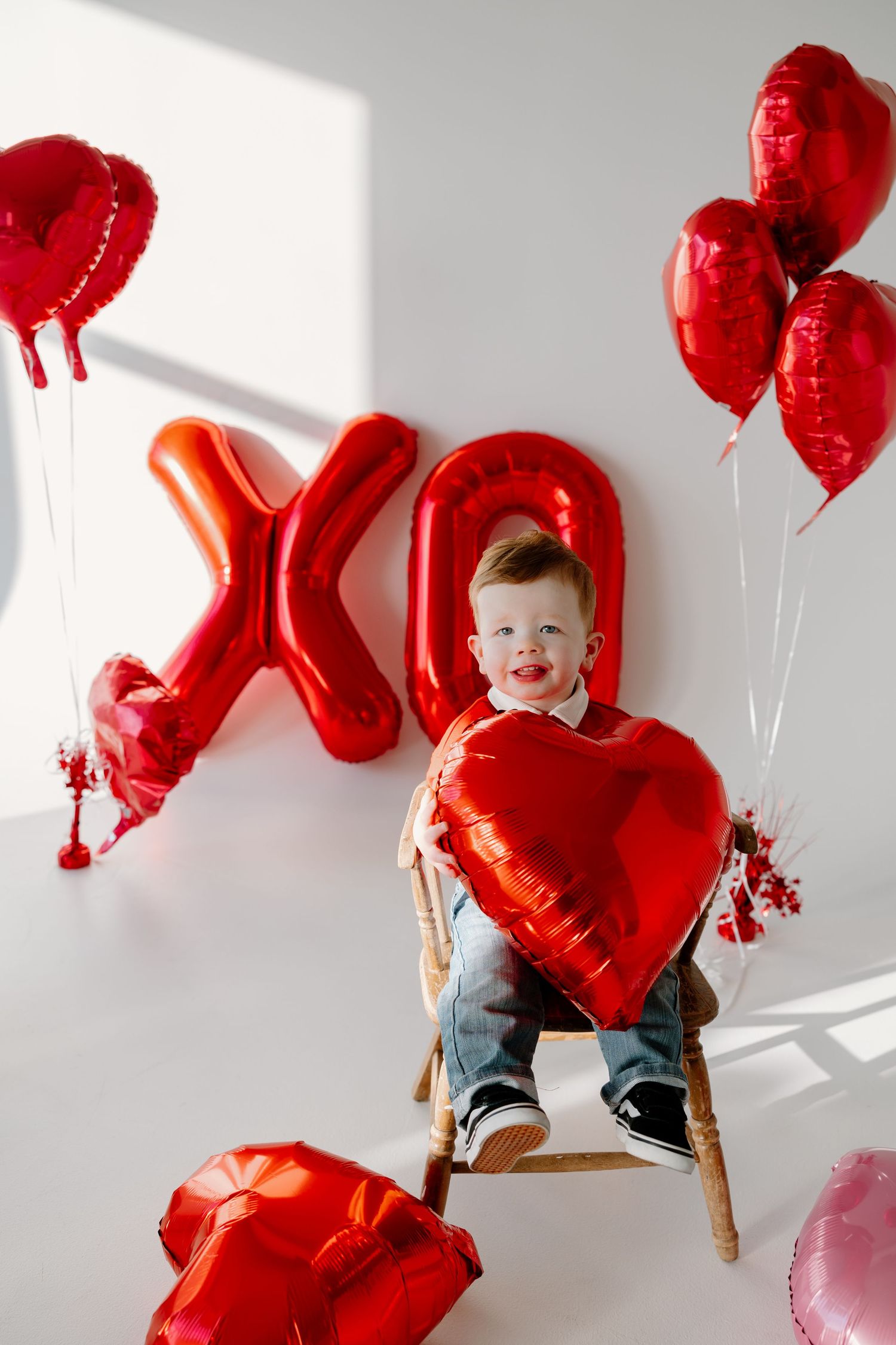 Festive Valentine's Day scene with red heart and XO letter balloons, wooden chair and natural lighting against white wall.