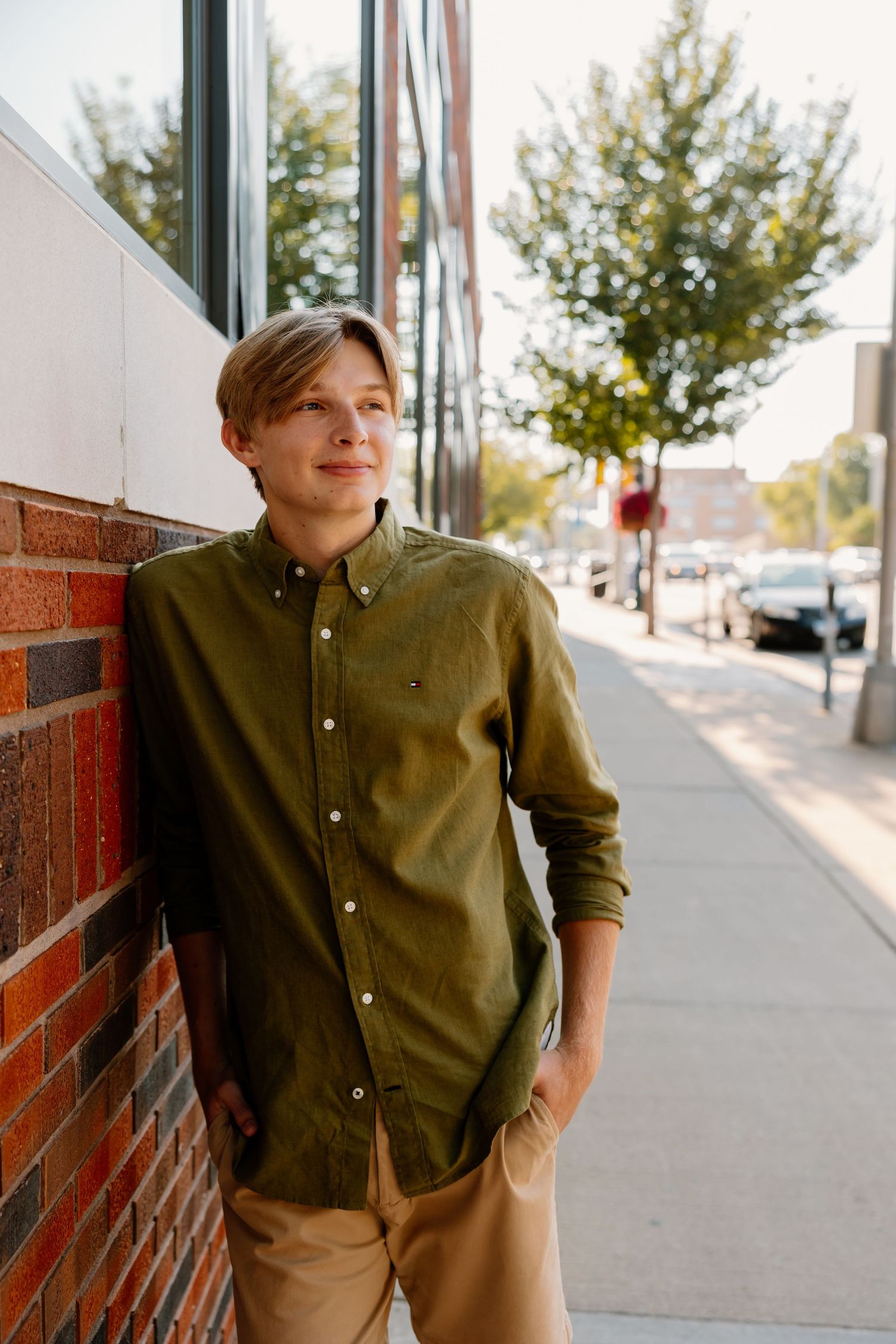 Person in olive button-down shirt poses casually against brick wall on sunny sidewalk.