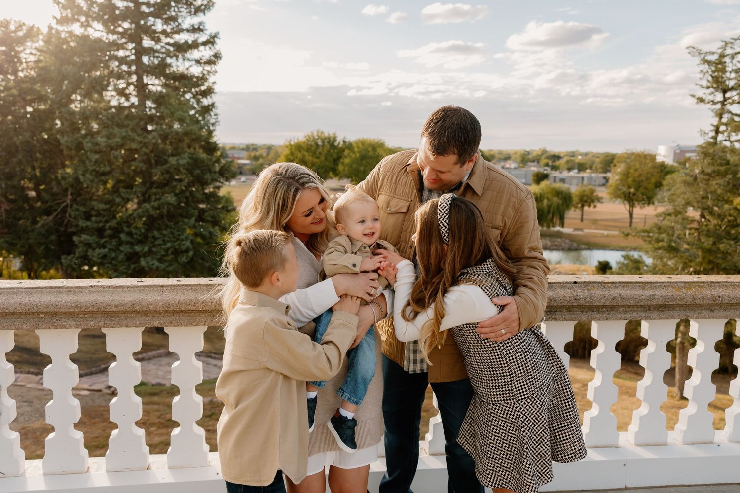Family of five sharing a warm embrace on a sunlit outdoor balcony surrounded by lush green trees.