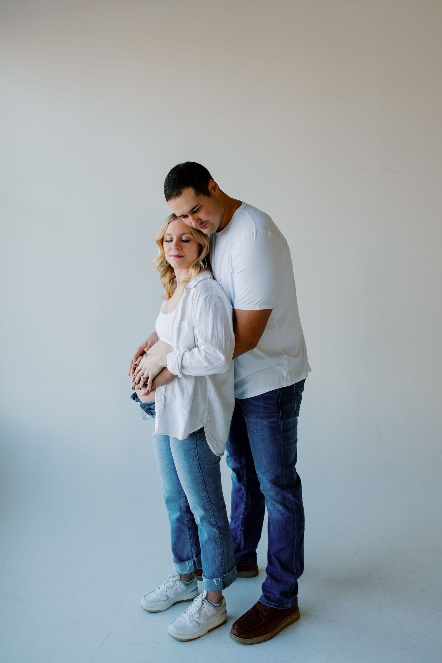 A couple embraces in a minimalist setting wearing casual attire during a maternity photoshoot. Riley Ro Photo Sioux Falls, SD