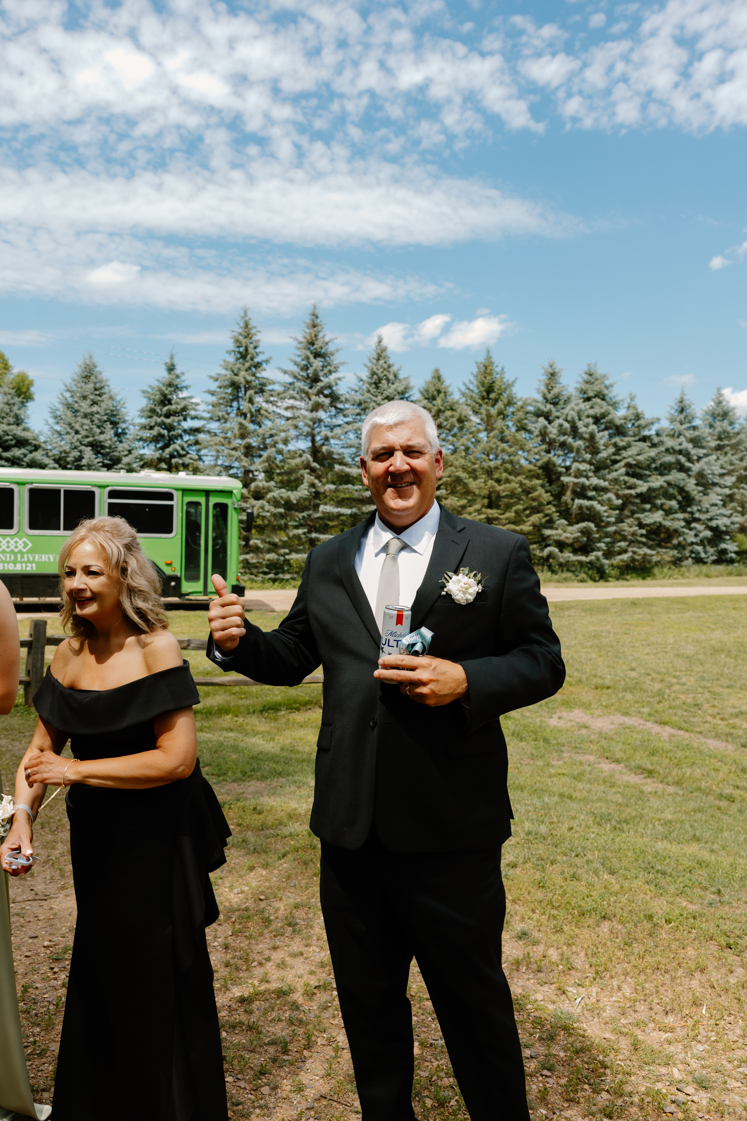 A smiling man in a black suit with boutonniere and woman in black dress at an outdoor wedding celebration.