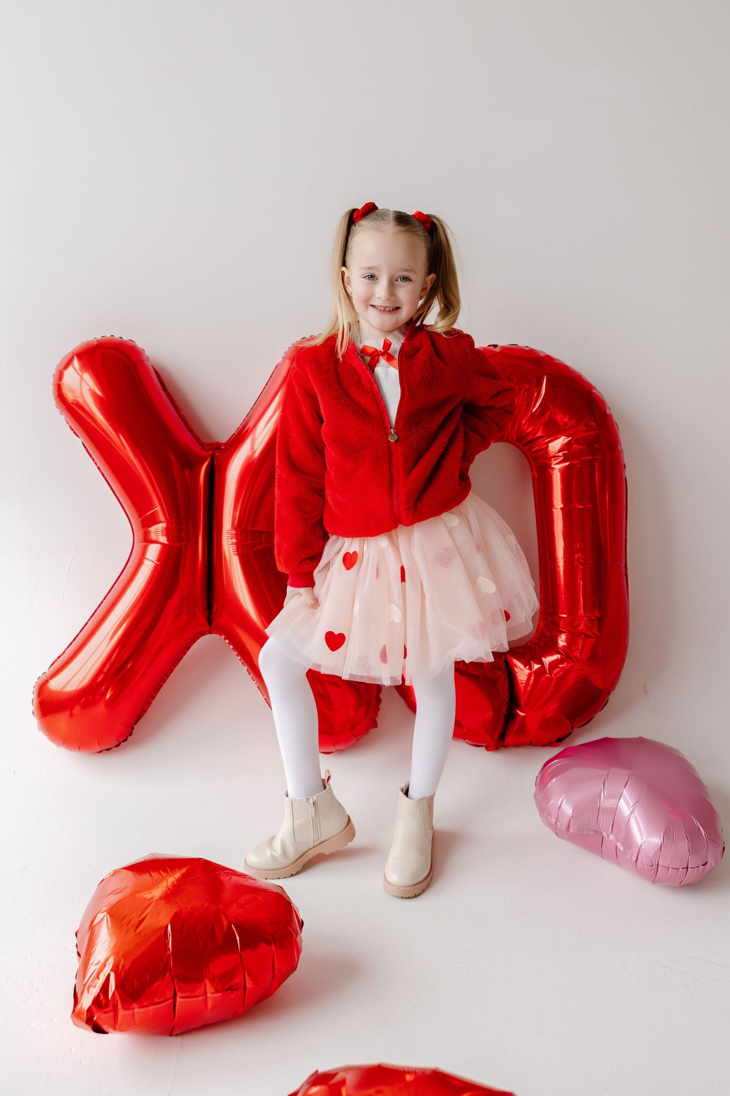 Valentine's Day setup with red balloon letters, heart decorations, and white tulle skirt with heart pattern against white wall.