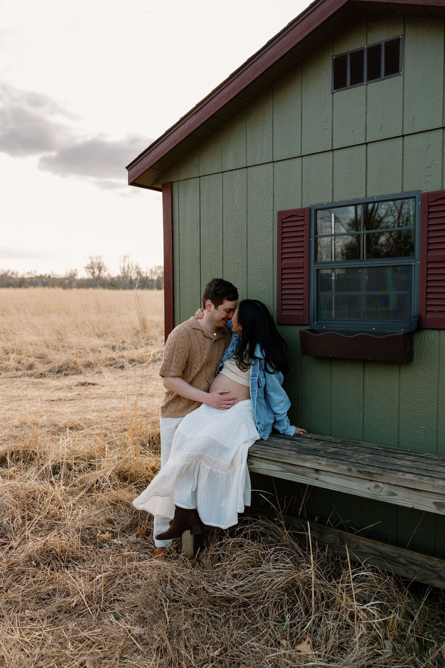 A romantic maternity photo of a couple sitting on a rustic green cabin's porch during sunset.