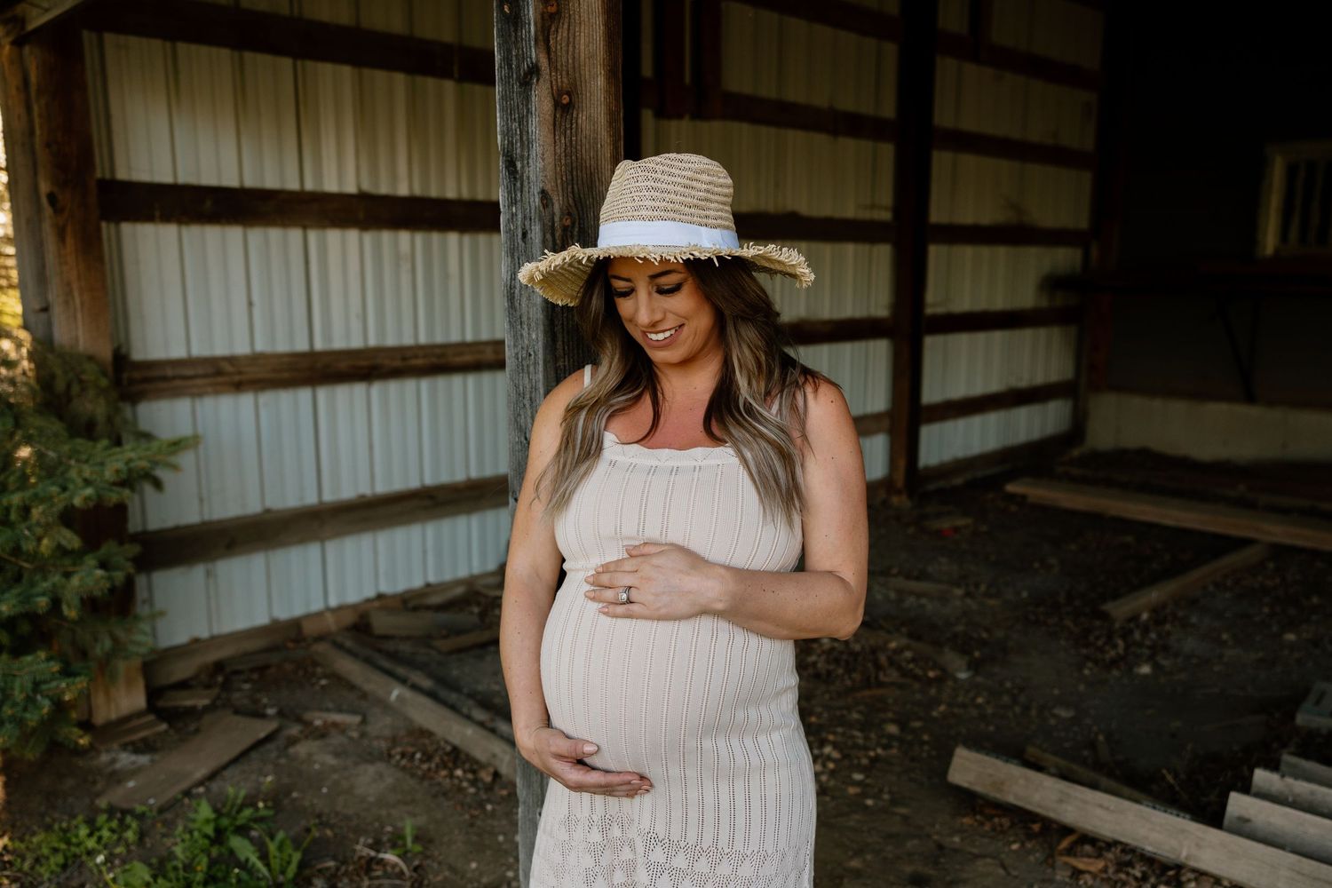 Expectant woman in white dress and sun hat smiles while cradling her baby bump in rustic barn setting.