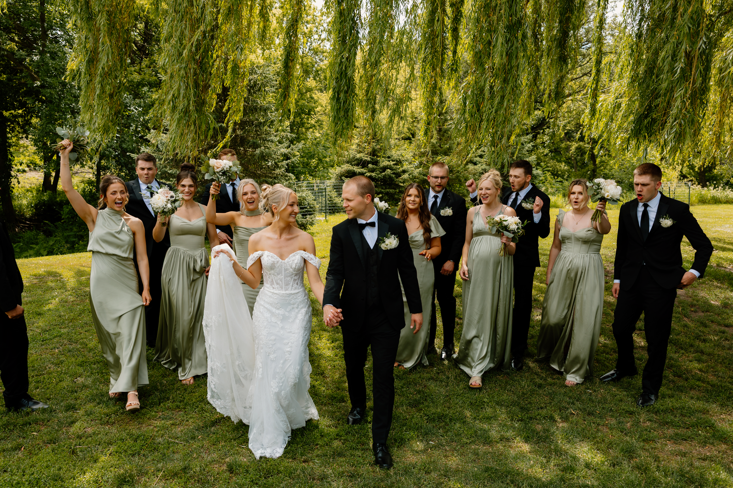 Wedding party walks outdoors under weeping willows, bride in white gown with groom and bridesmaids in sage green dresses.