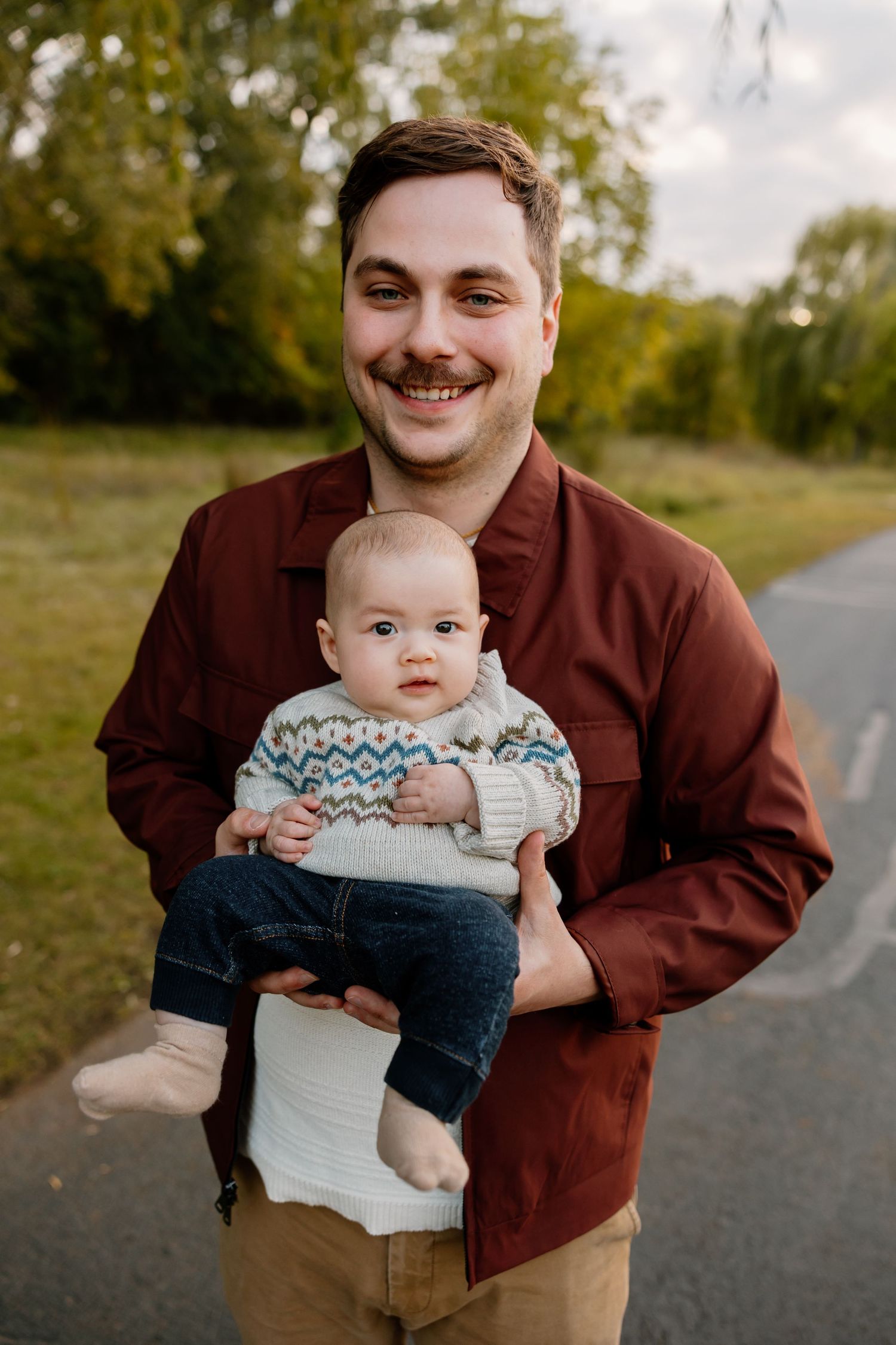 A series of four photos showing a parent in a brown jacket holding and playing with a baby wearing a patterned sweater outdoors. Riley Ro Ph