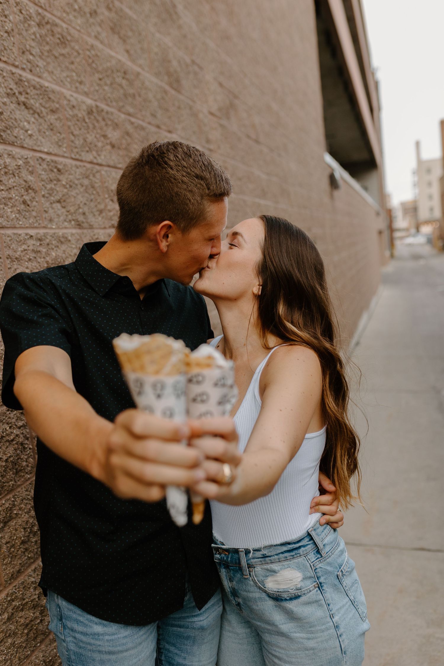 A couple sharing a kiss while holding ice cream against an urban wall.