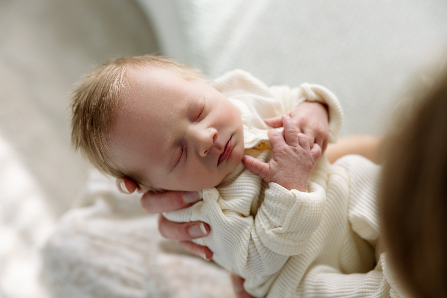 Peaceful sleeping newborn baby in cream outfit cradled gently in mother's hands, soft natural light photography.