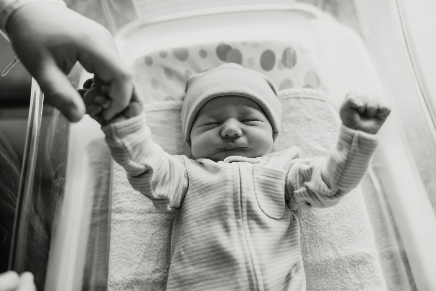 Black and white photo of a newborn baby stretching arms wide in a hospital bassinet, eyes closed peacefully.