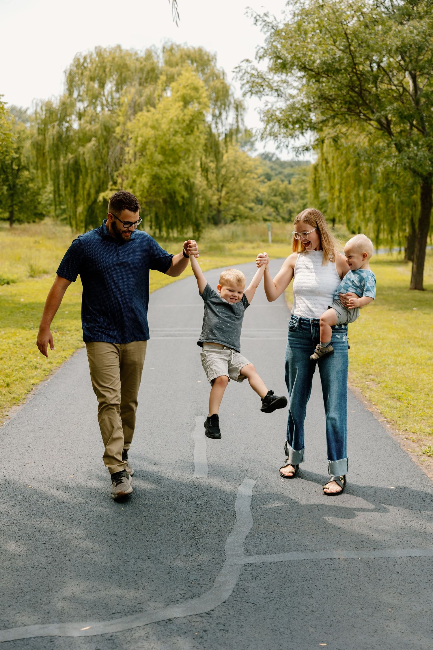 A joyful family enjoys a walk together on a path surrounded by willow trees in a sunny park setting.