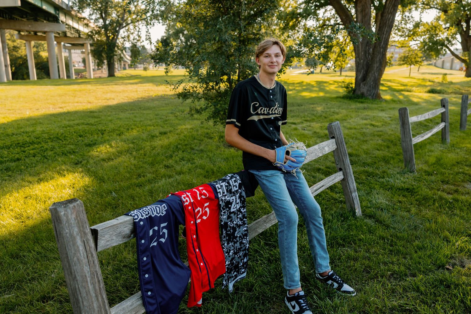 A young athlete sits casually on a wooden fence holding baseball gear against a sunlit grassy field background.