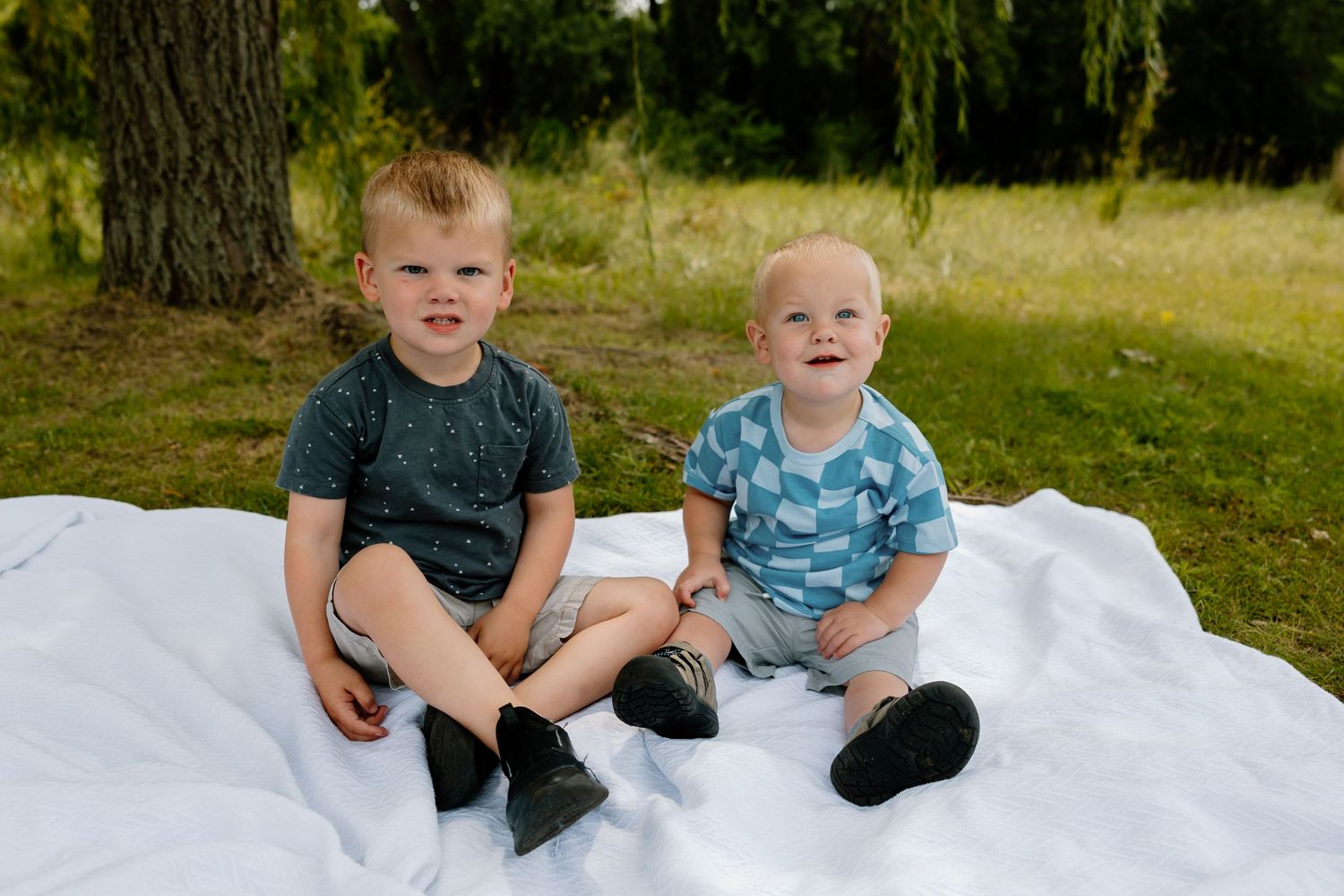 Young siblings giggle and play together on a blanket in a sunny outdoor setting.