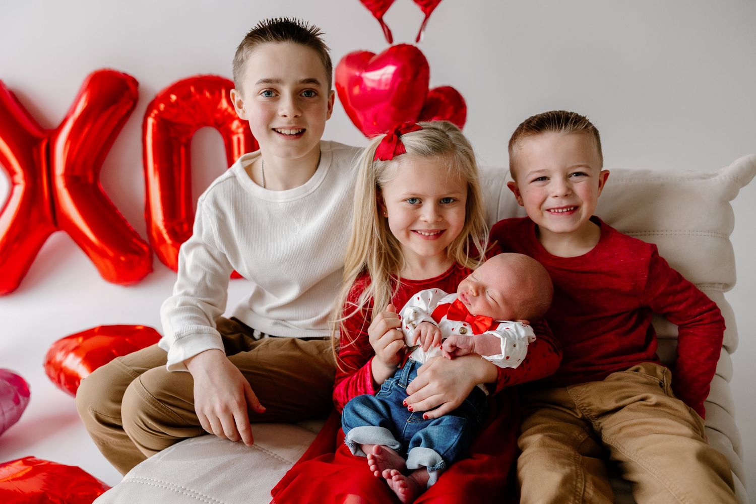 Children sit together on white couch with red heart balloons decorating wall for Valentine's Day photo session.