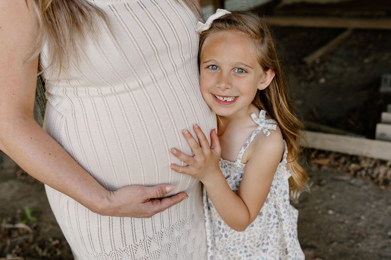 A young girl in a white ruffled dress gently touching a pregnant woman's belly in a tender maternity moment.