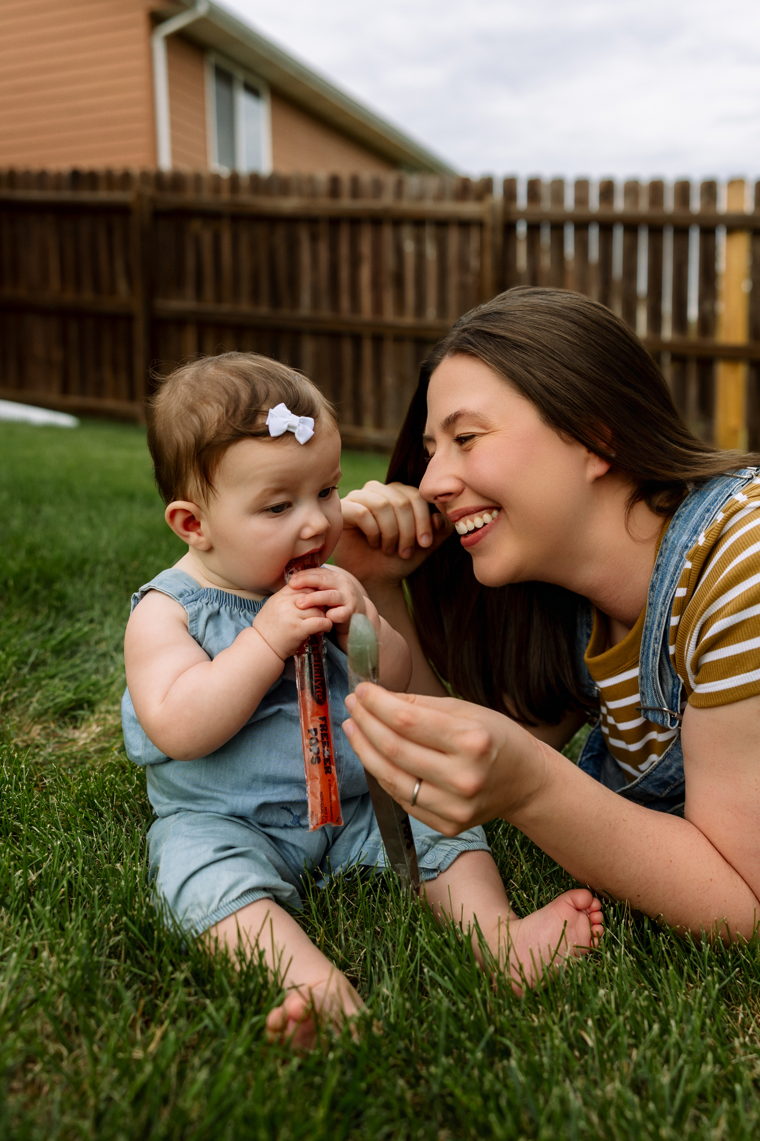 A smiling mother lies on green grass playing with her happy baby girl wearing a blue romper and bow.
