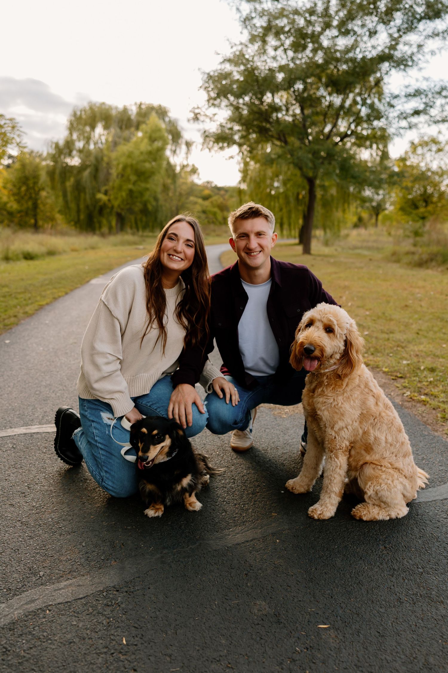 A couple sits on a path with their two dogs, a small dark one and larger goldendoodle, on an autumn evening. Riley Ro Photo Sioux Falls, SD