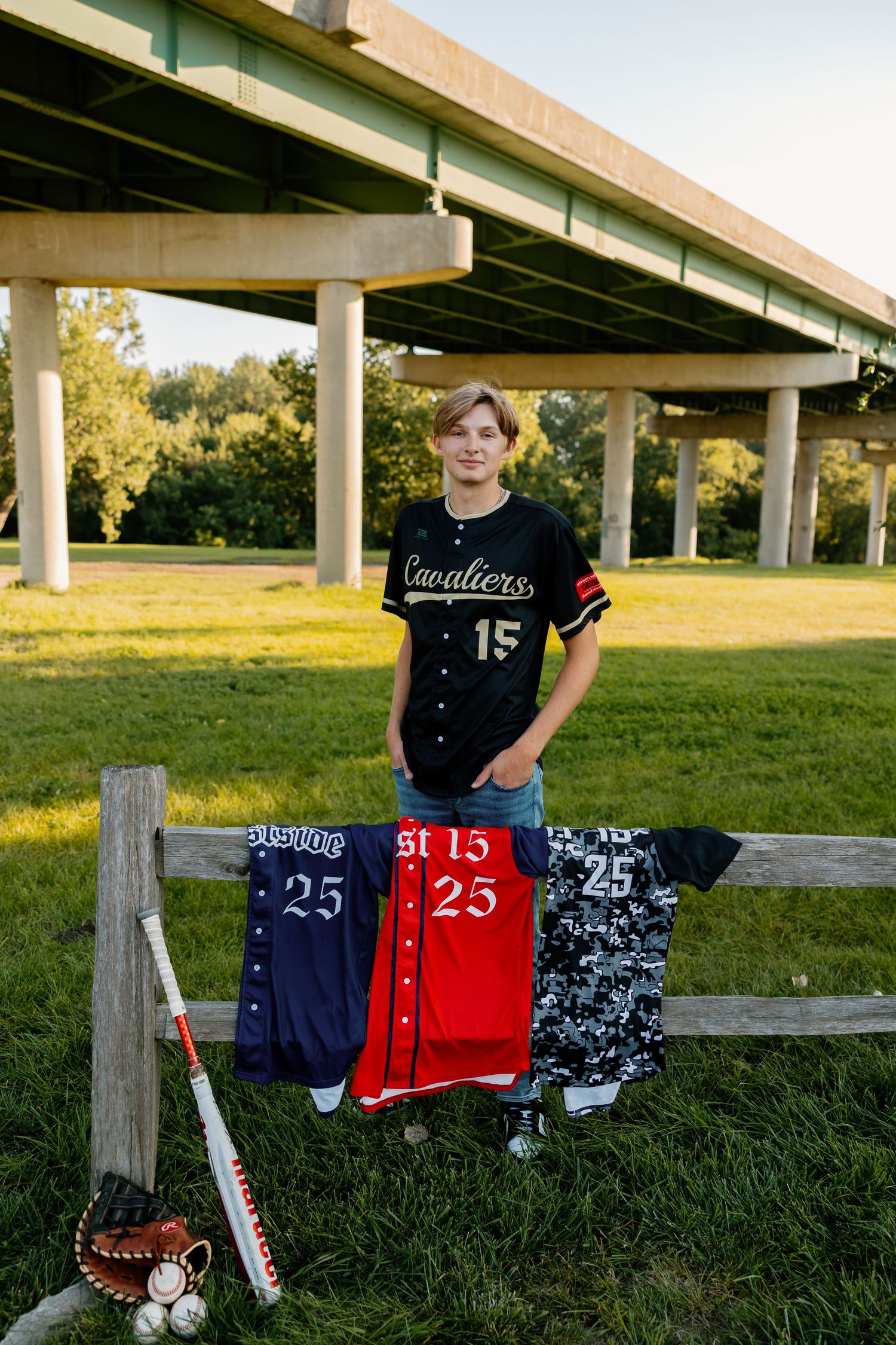 Baseball player with multiple jerseys and equipment displayed on fence under overpass.