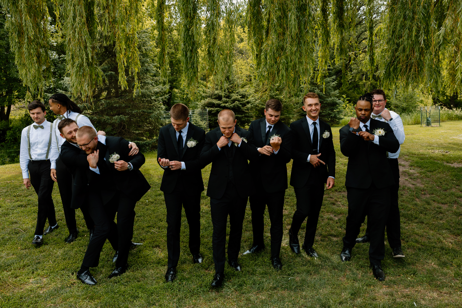 Groomsmen in black tuxedos posing playfully outdoors under weeping willow trees at a wedding.
