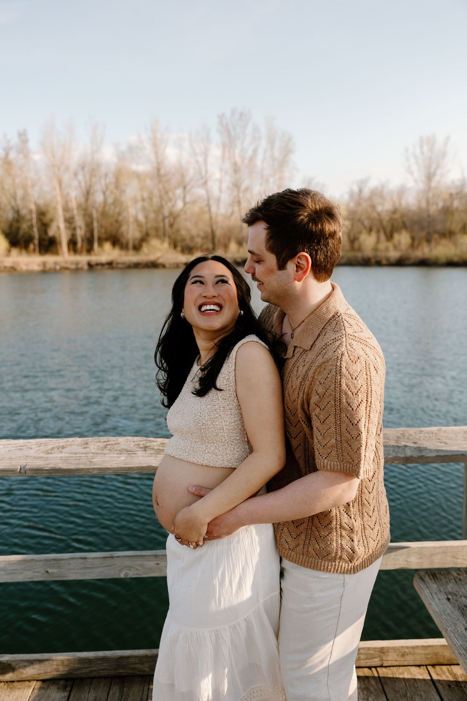 A joyful maternity photoshoot of a couple embracing on a wooden dock by a serene lake during sunset.