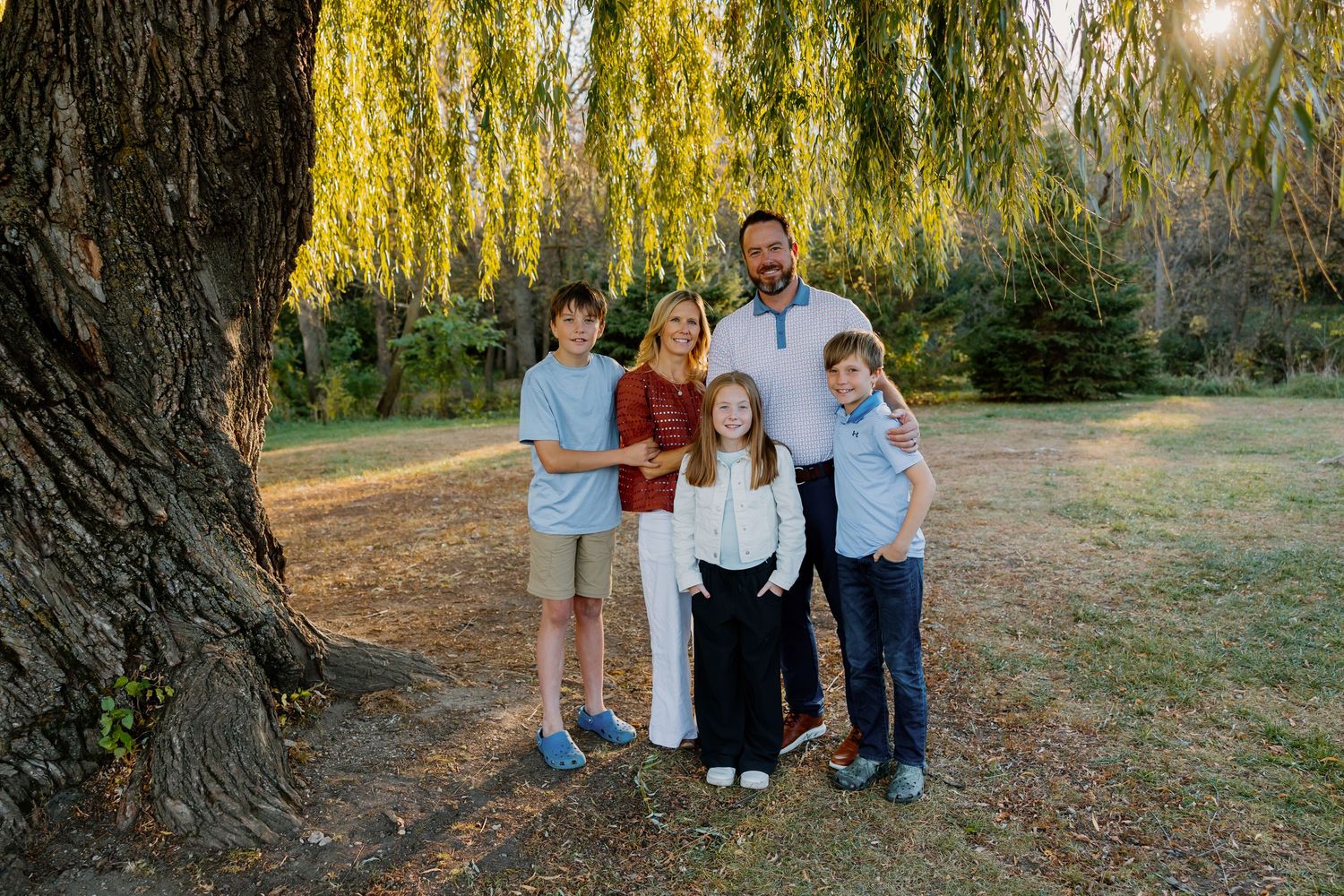 Family of five posing outdoors near a large willow tree in a sunny park setting.